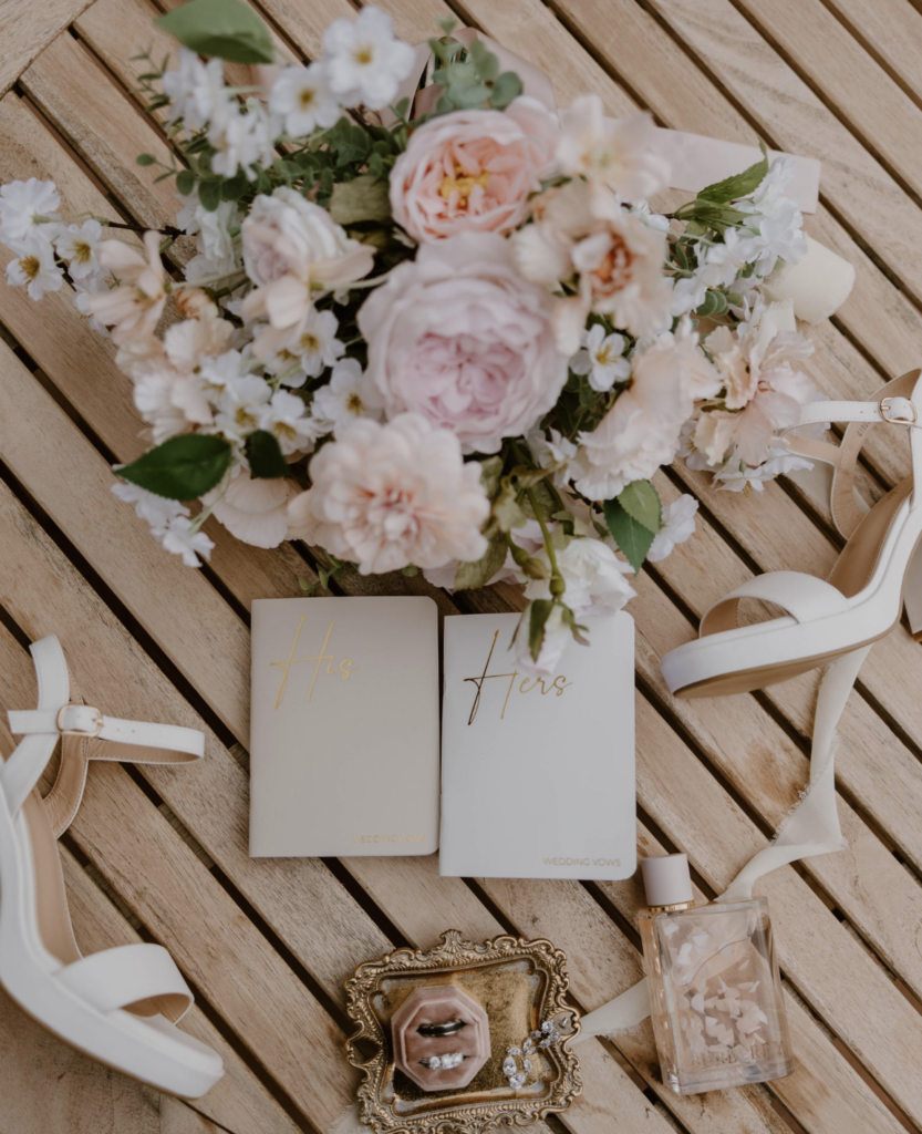 Custom flat lay with wedding day florals with his and hers vow books, wedding rings and perfume.